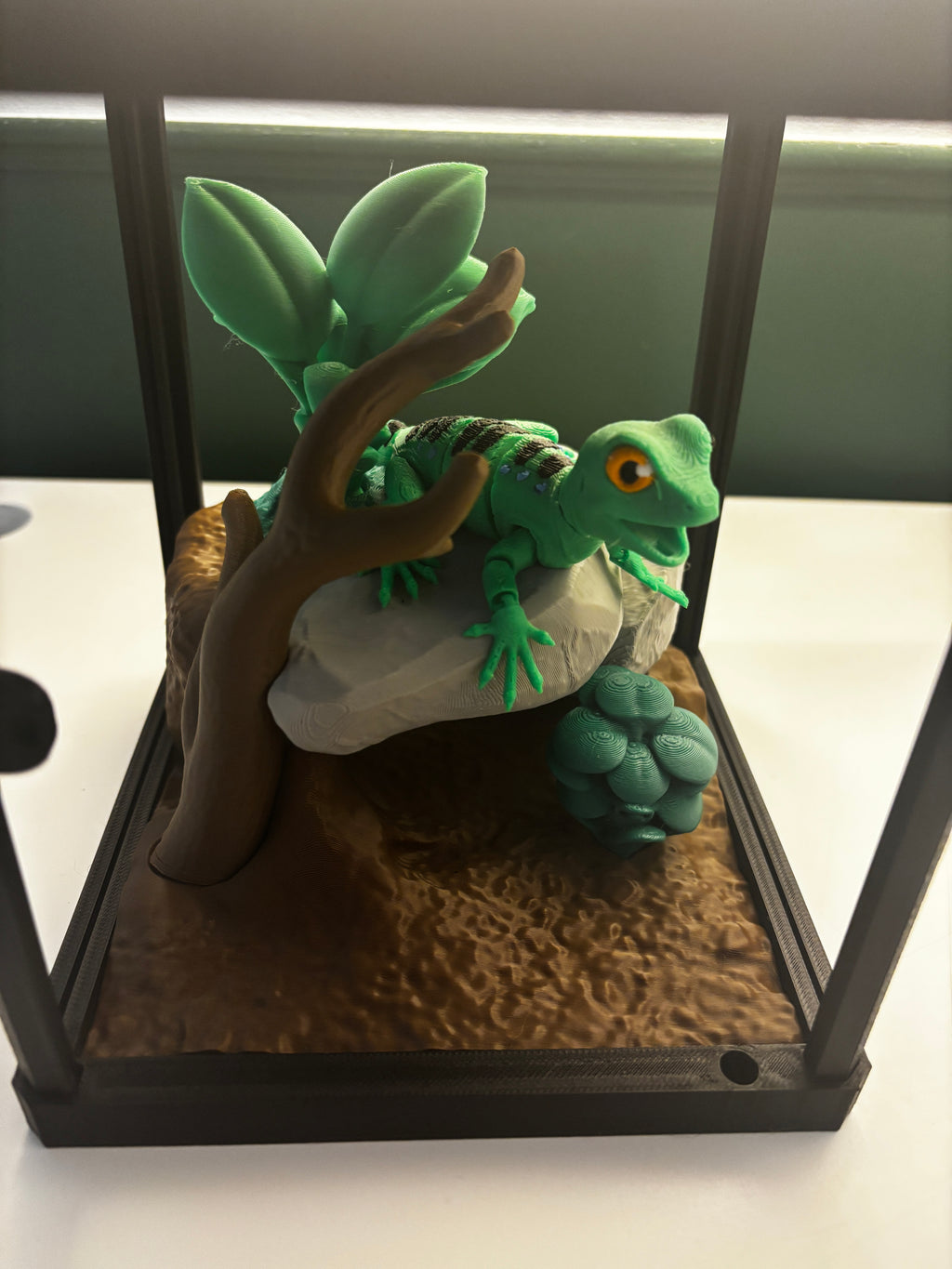 A 3D-printed articulated green iguana toy displayed inside a glass terrarium with a brown faux-wood branch, gray rock, textured brown substrate, and green plant leaves.