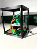 A black glass terrarium with a wooden branch and faux rocks, containing two green gecko figurines on a white surface against a dark green and white background.