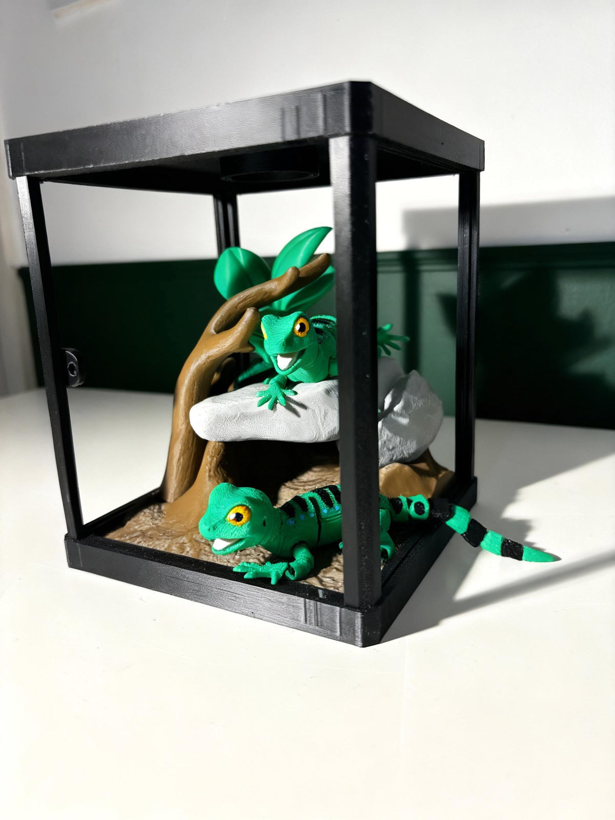 A black glass terrarium with a wooden branch and faux rocks, containing two green gecko figurines on a white surface against a dark green and white background.